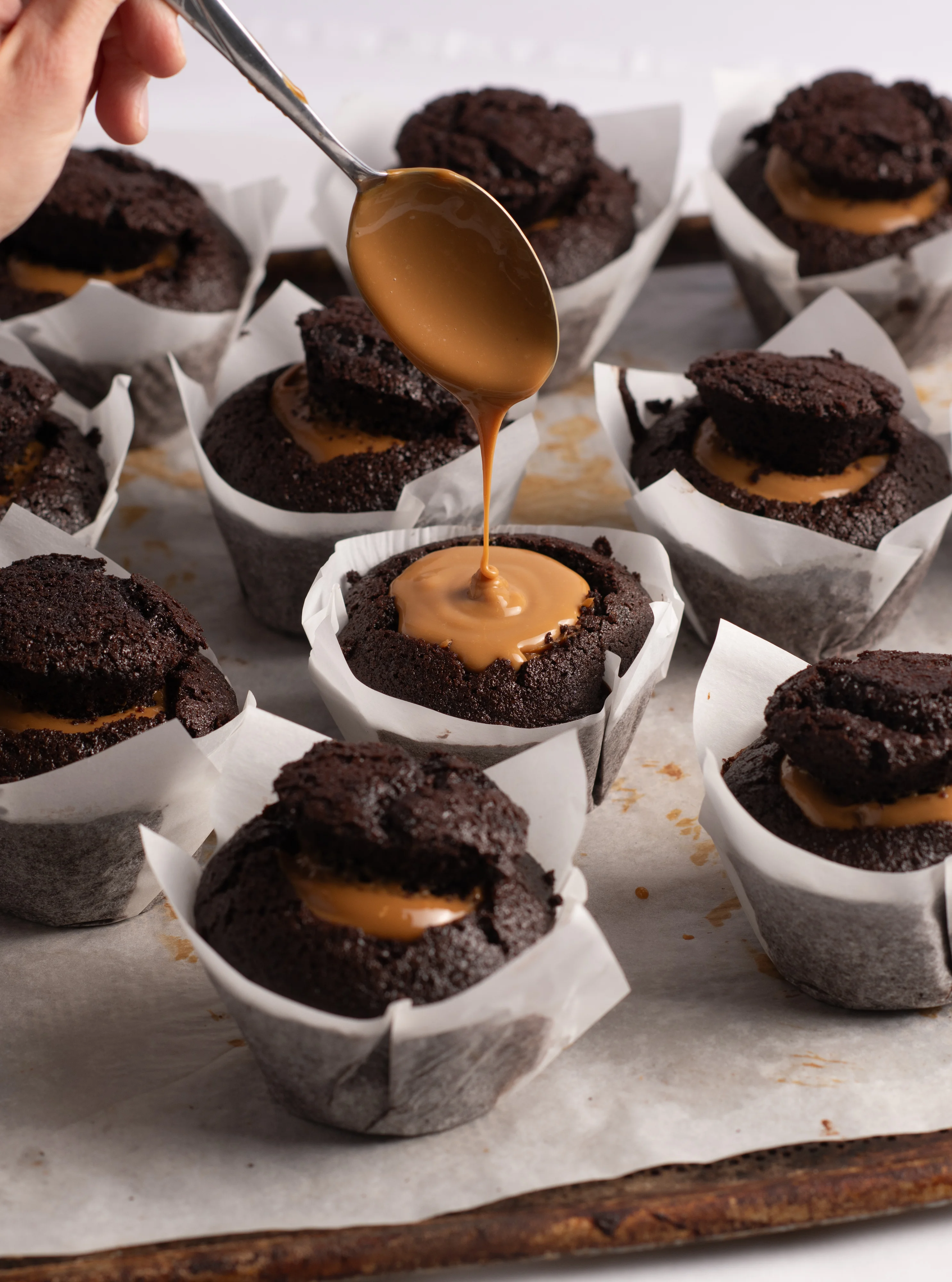 Warm salted caramel being spooned into the centre of a chocolate muffin, a tray of muffins in parchment liners around it.